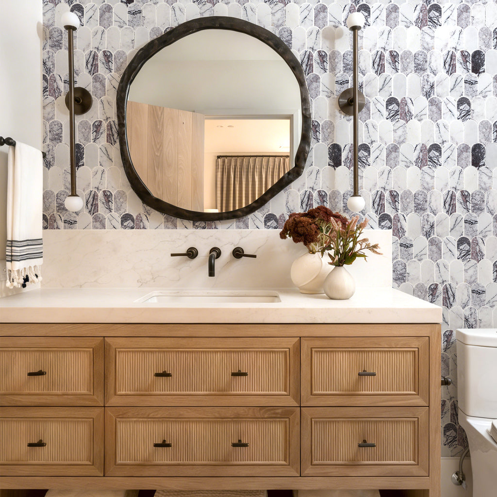 Bathroom vanity with round mirror, floral arrangement, and patterned wallpaper.