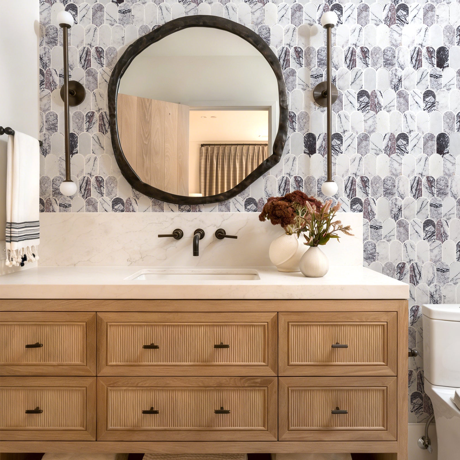Bathroom vanity with round mirror, floral arrangement, and patterned wallpaper.